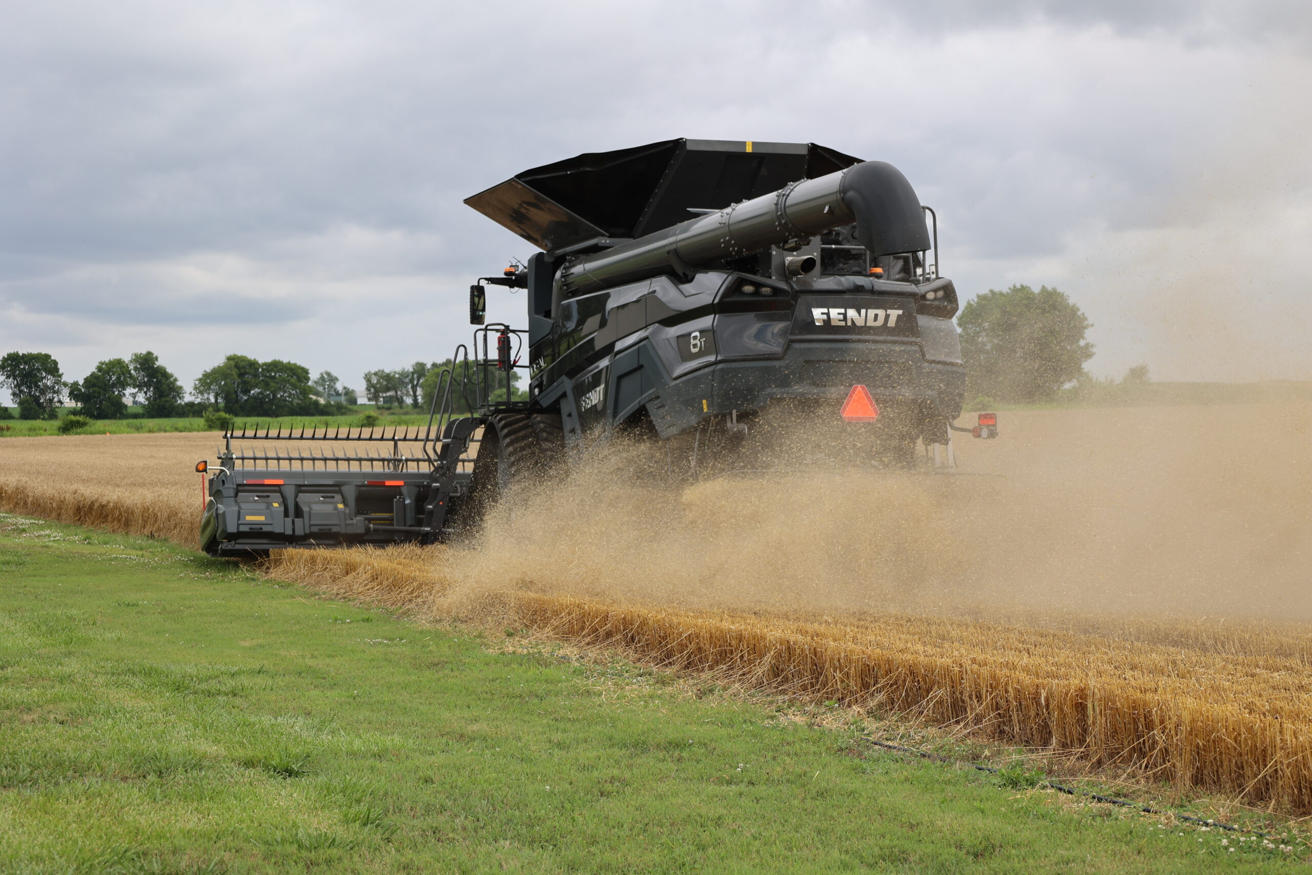 Fendt IDEAL combine harvesting wheat with rear view of grain discharge and chopper in action on a cloudy summer day.