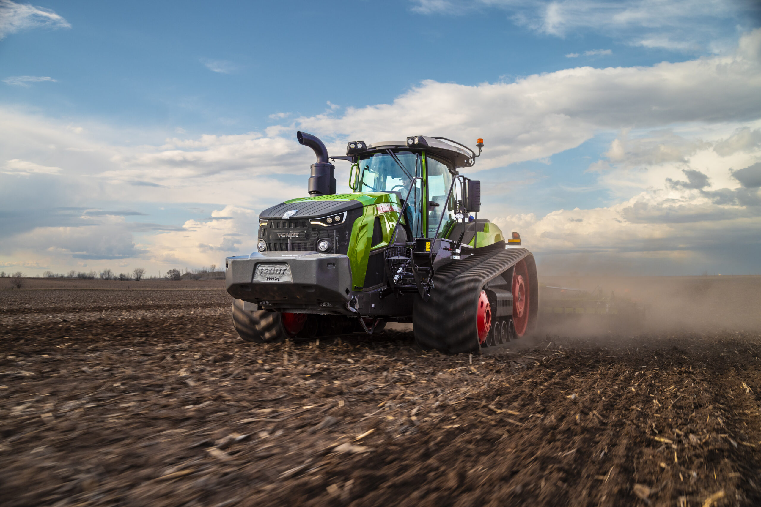 Fendt 1100 tracked tractor driving across a field, kicking up dust under a partly cloudy sky.