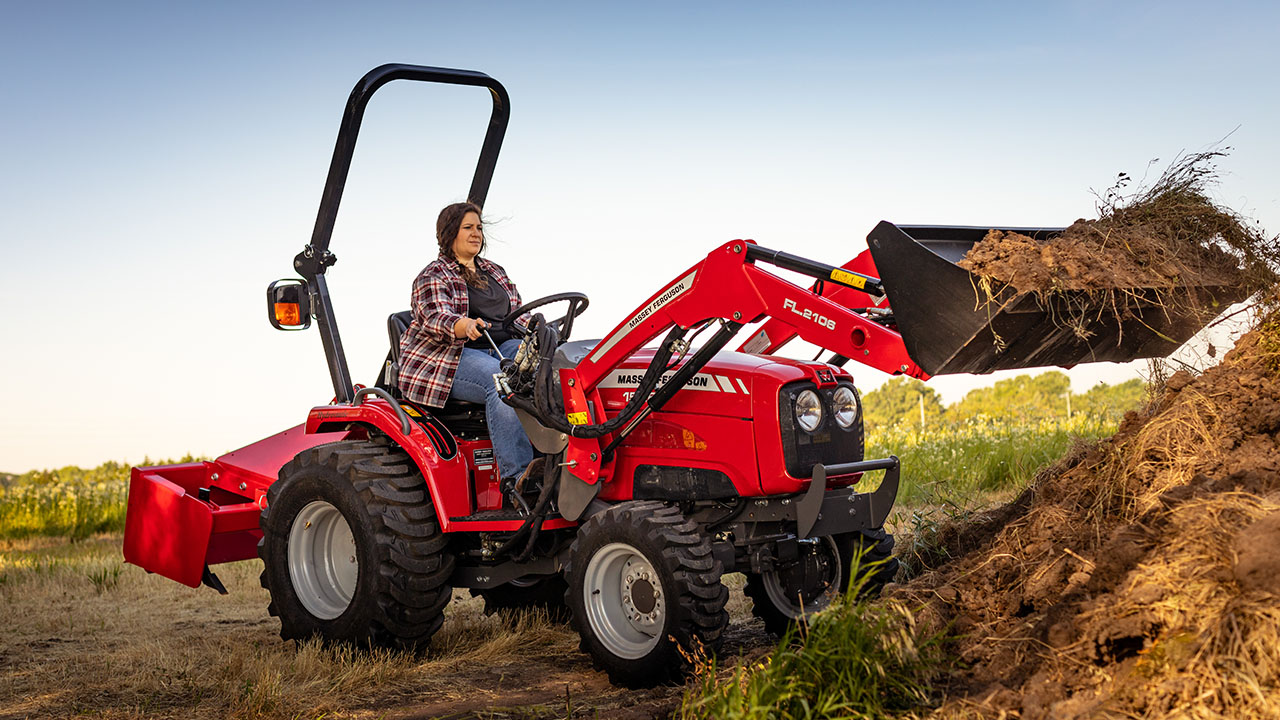 Woman operating Massey Ferguson 1526 compact tractor with front loader, moving dirt on a rural property.