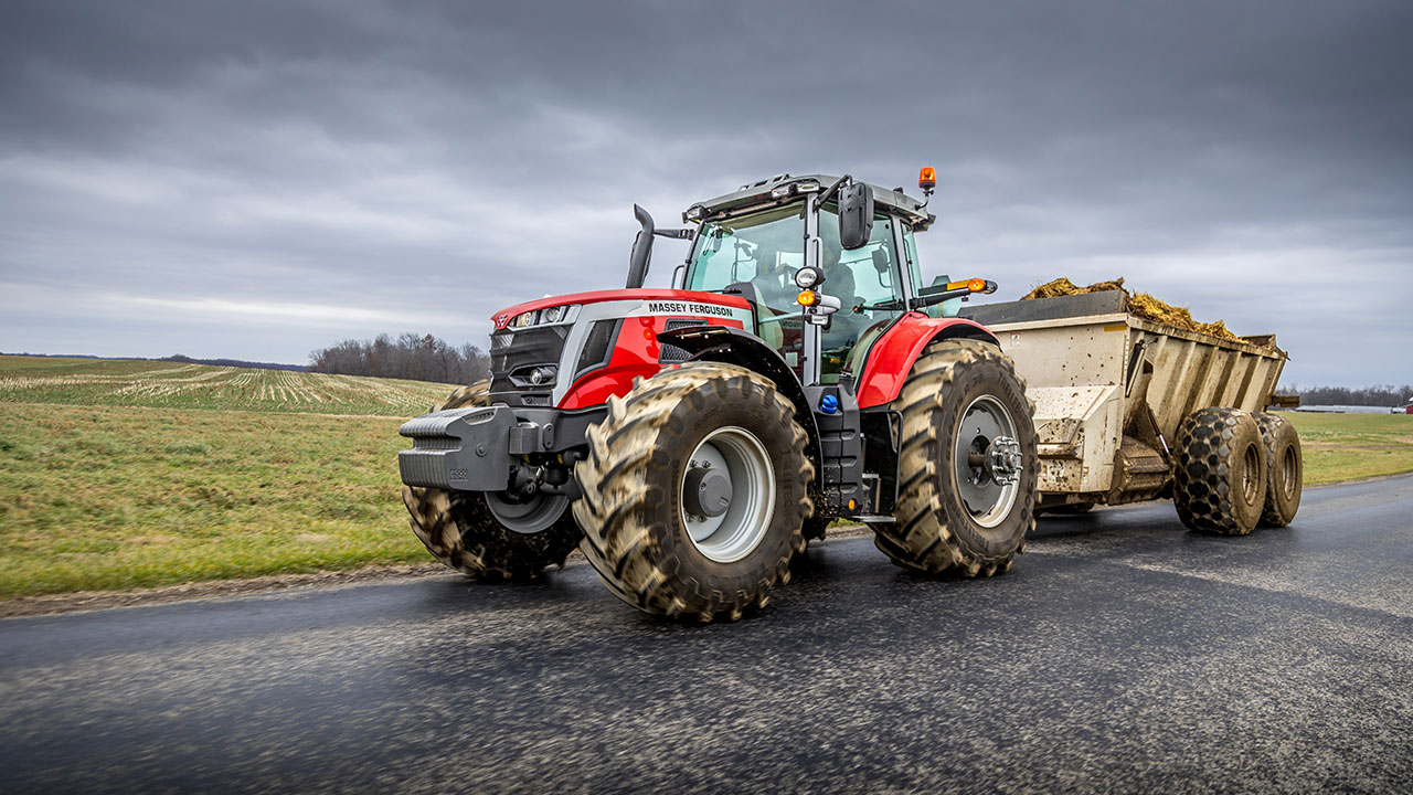 Massey Ferguson 7S Series tractor hauling a manure spreader on a rural road under a cloudy sky, built for heavy-duty farm work.