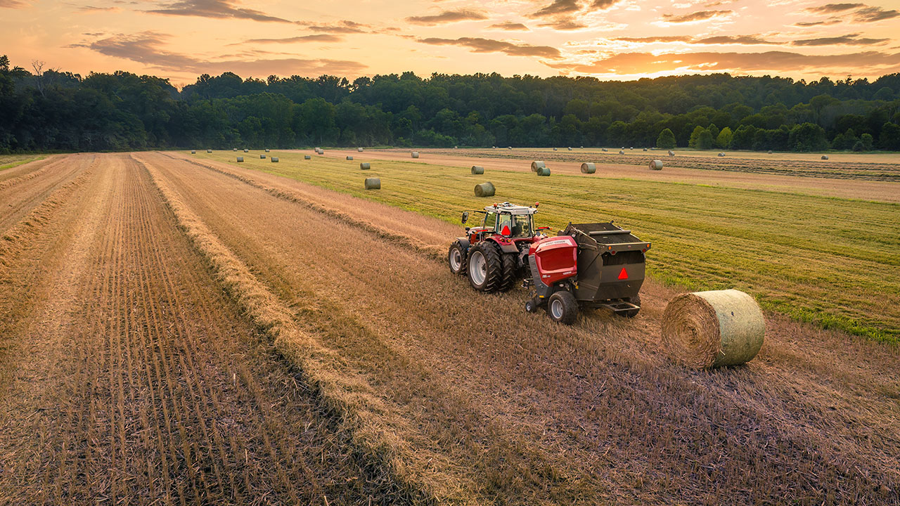 Hesston by Massey Ferguson 1 Series round baler releasing hay bale in a field at sunset with tractor.