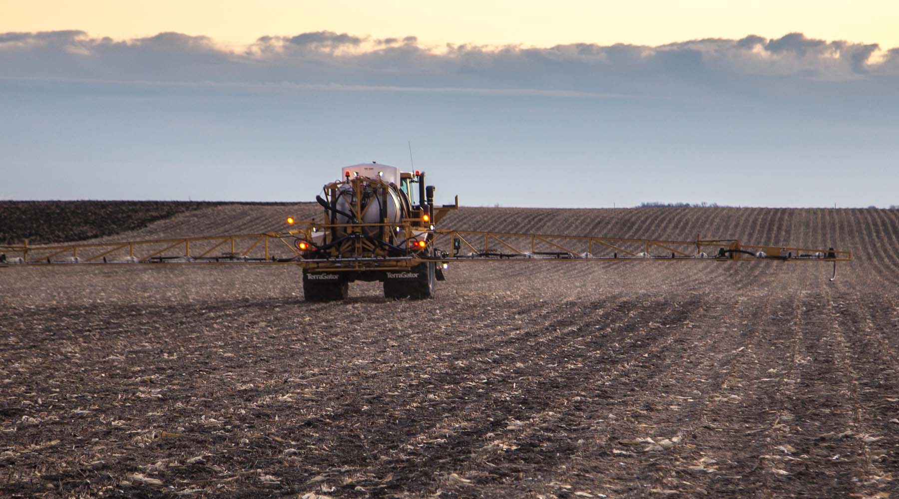 Rear view of TerraGator C Series sprayer with extended boom applying crop inputs across large, tilled field at dusk.
