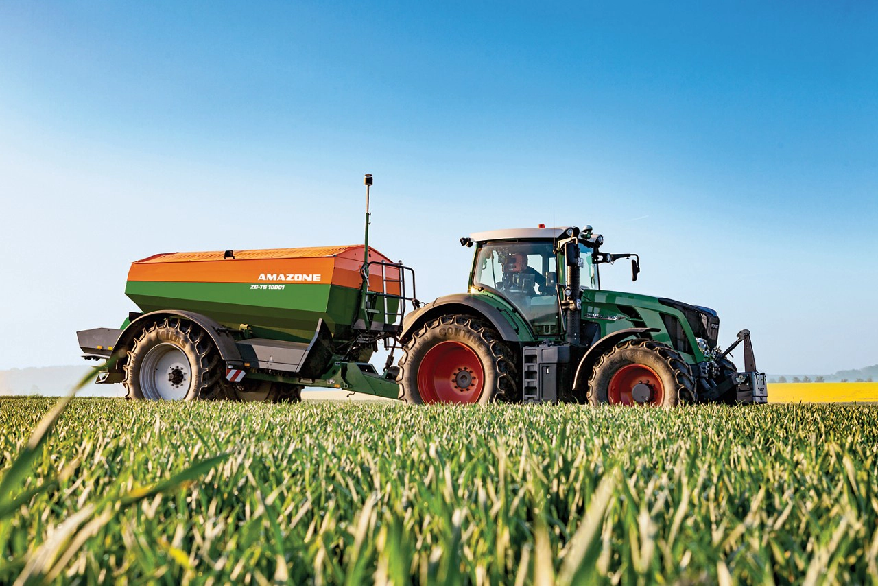 Fendt tractor towing an Amazone ZG-TS 10001 trailed fertilizer spreader in a lush green field under a clear blue sky, ideal for precision agriculture and efficient fertilizer application.