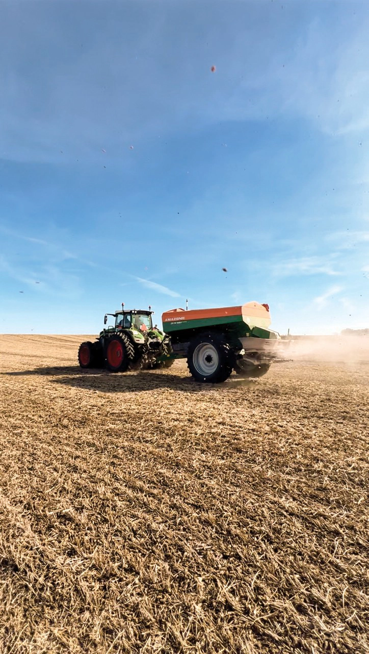 Fendt tractor towing an Amazone ZG-TS trailed fertilizer spreader applying nutrients in a harvested field under a clear blue sky, ensuring efficient soil preparation for the next planting season.
