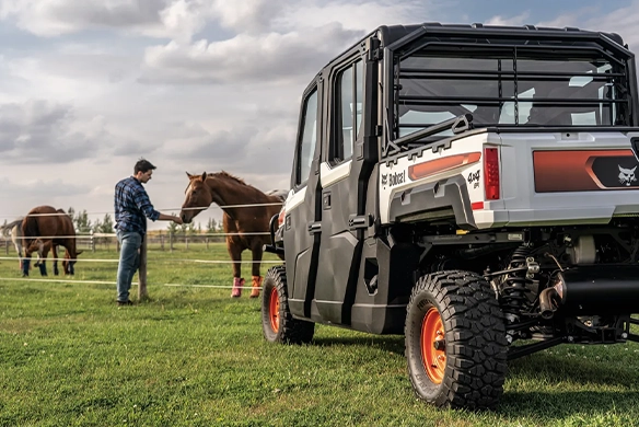 Bobcat UV34XL utility vehicle parked on a farm while a man feeds horses in a fenced pasture, demonstrating reliable 4x4 performance for daily agricultural chores.