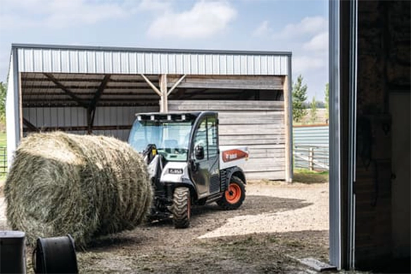Bobcat UW56 utility work machine equipped with a bale fork attachment, transporting a large round bale near a livestock shelter on a farm.