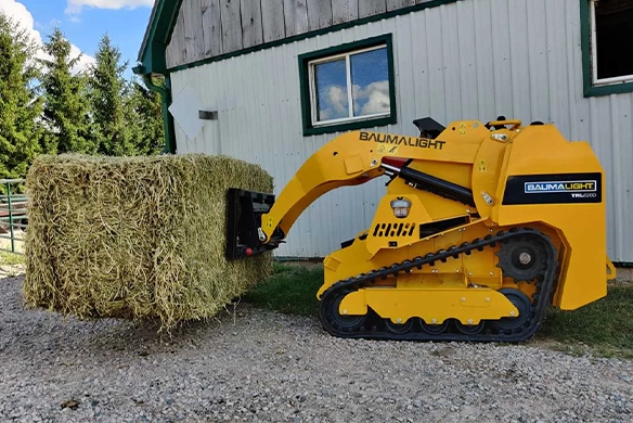 Compact Baumalight track loader transporting a large hay bale in front of a farm building.