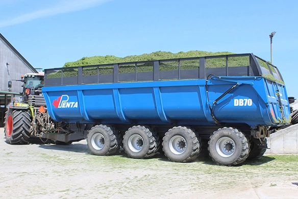 Blue Penta DB70 dump box trailer loaded with chopped forage, attached to a tractor outside a farm building.