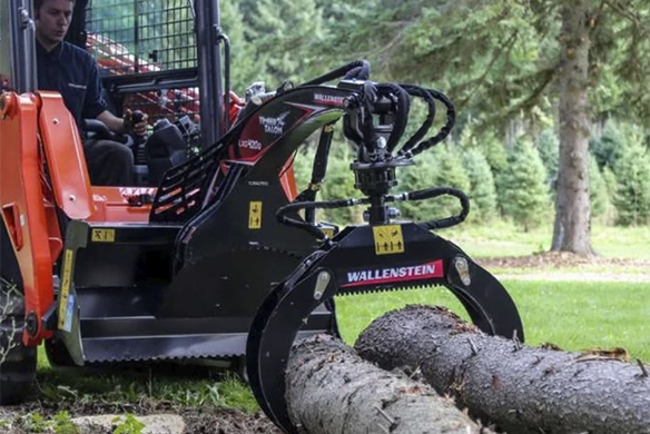 Close-up of Wallenstein tree grapple attachment mounted on a skid steer, grabbing cut logs in a wooded area
