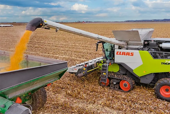 CLAAS TRION 740 combine with tracks unloading corn into a Brent grain cart, showcasing high-capacity grain handling in a cornfield.