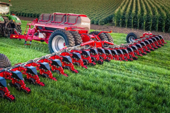 Wide view of a HORSCH Maestro SV planter with extended row units planting in a lush green field, paired with a Fendt tractor near tall corn crops.