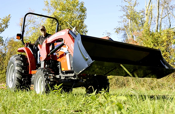Low-angle side view of a Massey Ferguson 2E Series compact tractor lifting the front loader bucket, highlighting ground clearance, loader strength, and efficiency for landscaping and loader work.