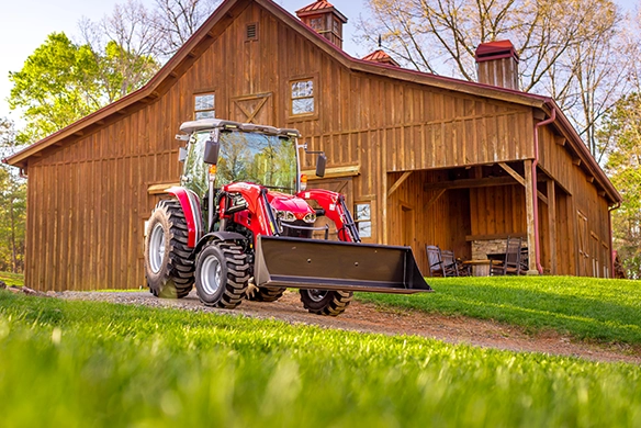 Massey Ferguson 2M Series compact tractor with front loader bucket in front of a wooden barn.