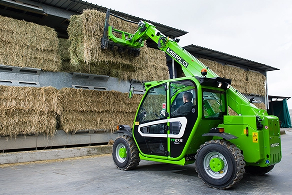 Merlo TF27.6 Plus telehandler lifting and stacking hay bales in a covered barn.