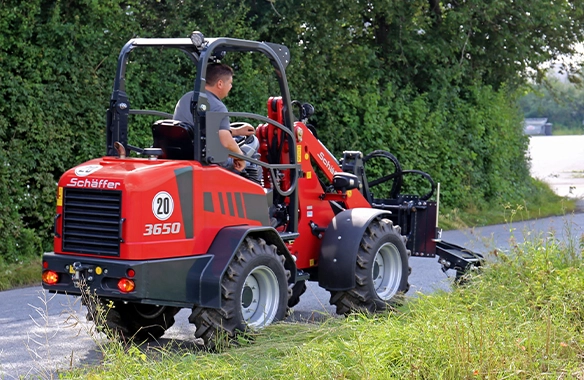 Rear view of Schäffer 3650 compact loader equipped with a flail mower trimming grass and brush along a rural roadside
