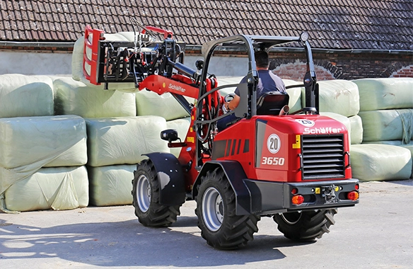 Schäffer 3650 compact loader moving wrapped hay bales with a bale grab in a storage yard.