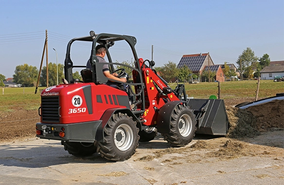 Schäffer 3650 compact loader using a general-purpose bucket to scoop silage from a feed pad on a sunny day.