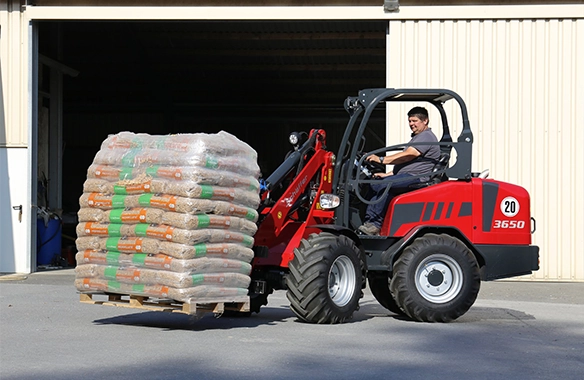 Schäffer 3650 loader carrying a full pallet of bagged materials into a warehouse with a pallet fork attachment.