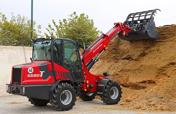 Schäffer 6680 T telescopic loader digging into a silage pile with a grapple bucket on a farm.
