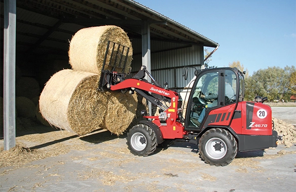 Schäffer 4670Z wheel loader stacking large round hay bales inside a barn using a bale fork attachment.