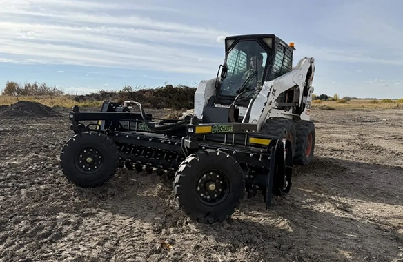 Schulte SV1000 rock picker attachment connected to a skid steer, operating on rough terrain for efficient rock and debris collection.