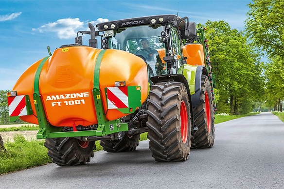 Front view of Claas tractor with an Amazone FT 1001 front tank system, driving along a tree-lined rural road.
