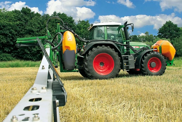 Side view of a Fendt tractor equipped with an Amazone UF-01 mounted sprayer and FT 1001 front tank, operating in a dry, harvested field with dense tree line in the background.