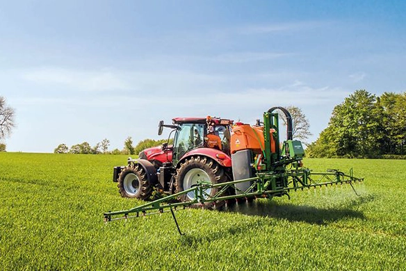 Red Massey Ferguson tractor operating an Amazone UF02 mounted sprayer in a green field with light trees in the background.