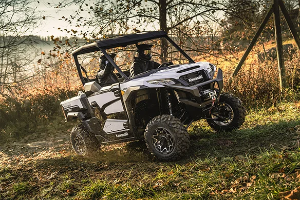 White Kawasaki Ridge Ranch navigates a grassy off-road trail in the woods during golden hour with two passengers.