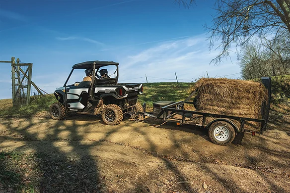 White Kawasaki Ridge Ranch towing a trailer loaded with hay bales up a dirt incline on a sunny farm.