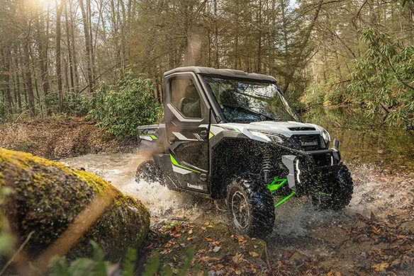 Black and green Kawasaki Ridge Ranch splashes through a shallow forest stream with enclosed cab and rugged tires.