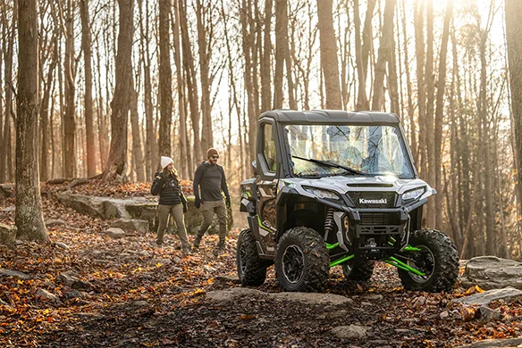 Black Kawasaki Ridge XR HVAC parked in a forest clearing as two hikers approach on a leaf-covered trail.