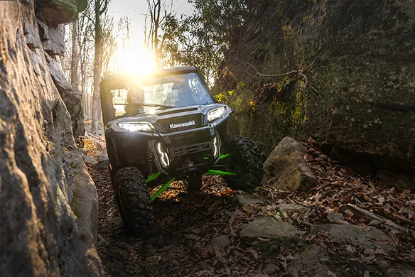 Black Kawasaki Ridge XR HVAC carefully maneuvers through a tight rocky forest trail at sunrise.