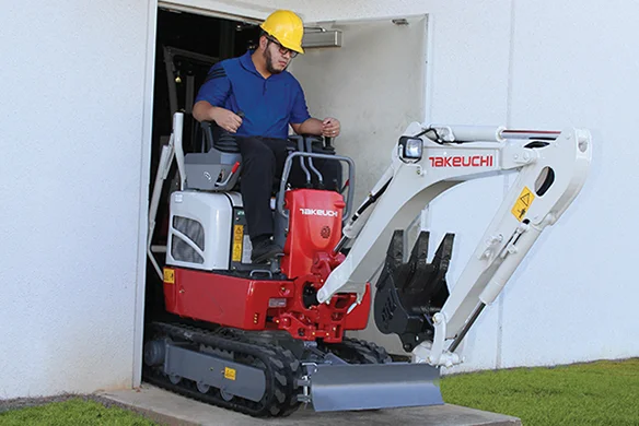 Worker operating Takeuchi TB210R compact excavator, exiting a building through a narrow doorway.