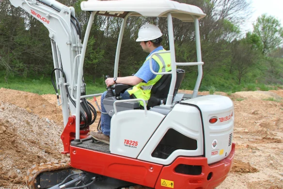 Takeuchi TB225 mini excavator lifting dirt on uneven terrain with operator in safety gear.