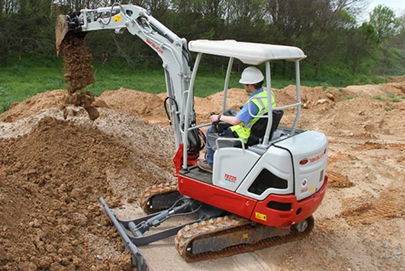 Takeuchi TB225 mini excavator lifting dirt on uneven terrain with operator in safety gear.