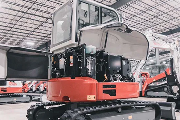 Takeuchi TB335R compact excavator with service panels open, showing engine and components in a warehouse setting.