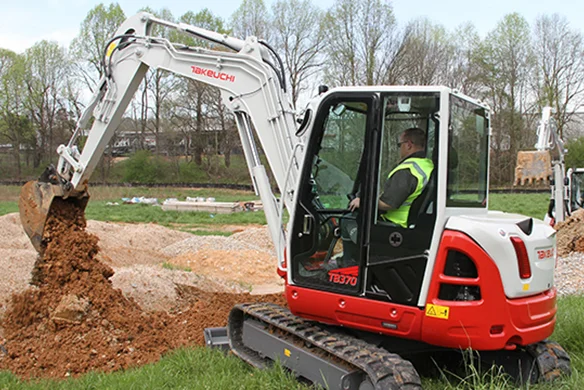 Takeuchi TB370 compact excavator lifting soil on a grassy jobsite, operated by worker in safety gear.