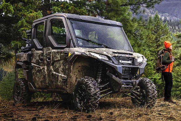 Kawasaki camo side-by-side utility vehicle parked in the forest with hunter standing nearby.