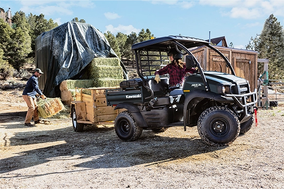 Black Kawasaki Mule SX towing a trailer loaded with hay while a farmer loads bales near a barn and haystack.