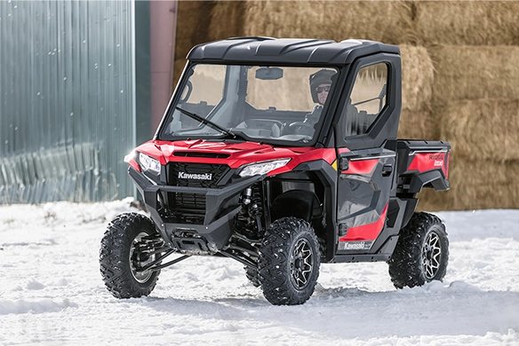Red Kawasaki Ridge HVAC drives through snow-covered terrain near a barn with the driver visible through the enclosed cab.