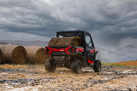 Rear view of a red Kawasaki Ridge HVAC hauling hay bales near a field under stormy skies.