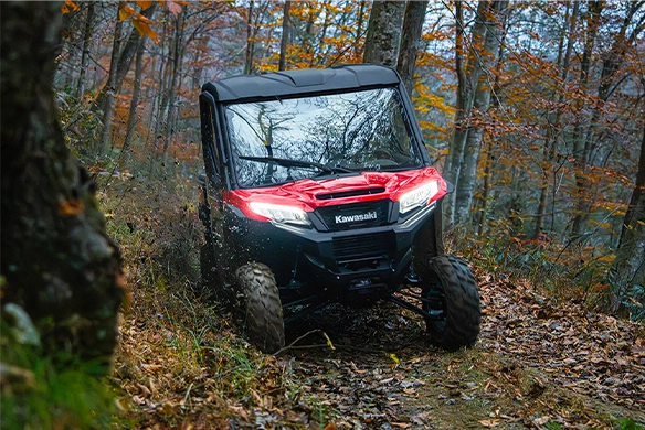 A red and black Kawasaki Ridge HVAC model driving through a forest trail in autumn, surrounded by fallen leaves and trees.