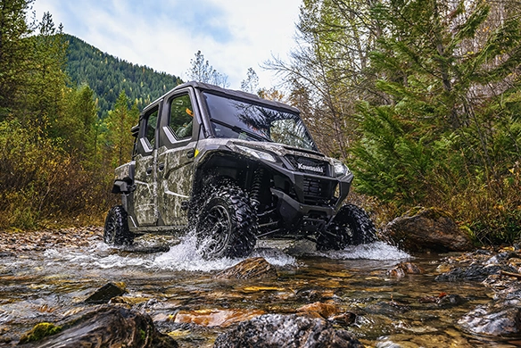 A Kawasaki Ridge CREW HVAC model in camo finish driving through a shallow rocky stream surrounded by trees.