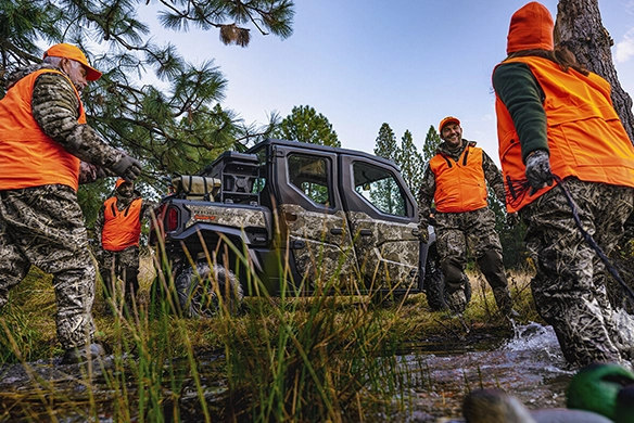 A group of hunters in camo and orange vests gathered near a Kawasaki Ridge CREW HVAC camo vehicle parked beside a creek in a wooded area.