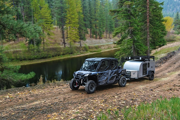 Kawasaki Ridge XR Crew Limited towing a small camping trailer through a wooded trail beside a peaceful river.