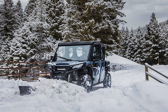 Kawasaki Ridge XR Crew Limited plowing snow in a forested winter scene, clearing a path with its front-mounted blade.