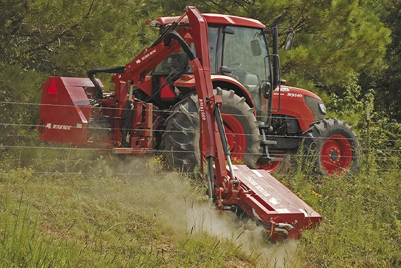 A red Bush Hog boom mower attached to a Mahindra tractor, cutting vegetation along a fence line.