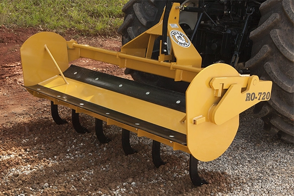 A yellow Bush Hog RO-720 landscape rake attached to a tractor, preparing gravel on a work site.