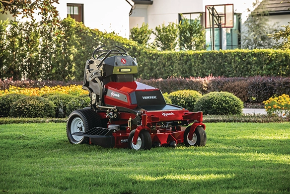 Exmark VERTEX stand-on mower parked on a freshly cut residential lawn with surrounding shrubs.